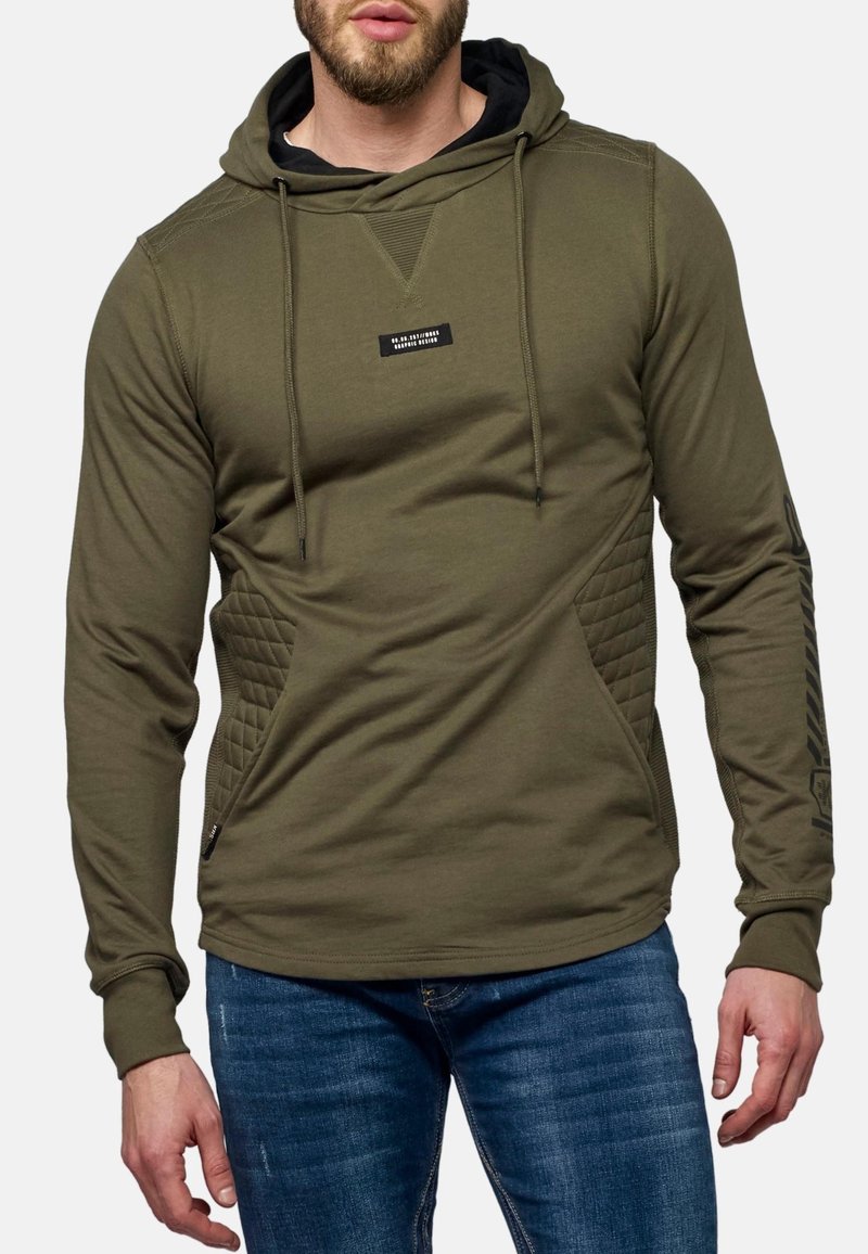 Uomo che indossa una felpa con cappuccio verde oliva con pannelli laterali trapuntati e jeans blu scuro, in piedi con le mani rilassate ai lati.