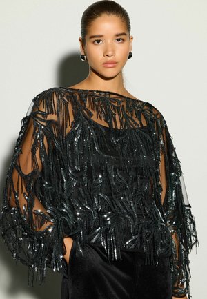 Joven con el cabello peinado hacia atrás lleva una blusa negra transparente adornada con flecos de cuentas negras brillantes y pantalones de terciopelo negro sobre un fondo liso.