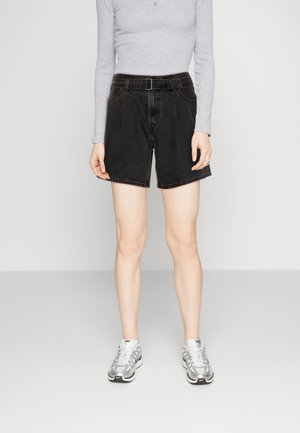 Persoon draagt zwarte denim shorts met een hoge taille en riem, grijs shirt met lange mouwen en zilveren Nike sneakers, staand tegen een effen achtergrond.