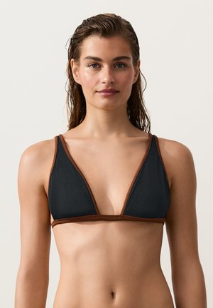 Kobieta z mokrymi włosami, ubrana w czarno-brązowy top bikini, zwrócona przodem, na jednolitym jasnym tle.