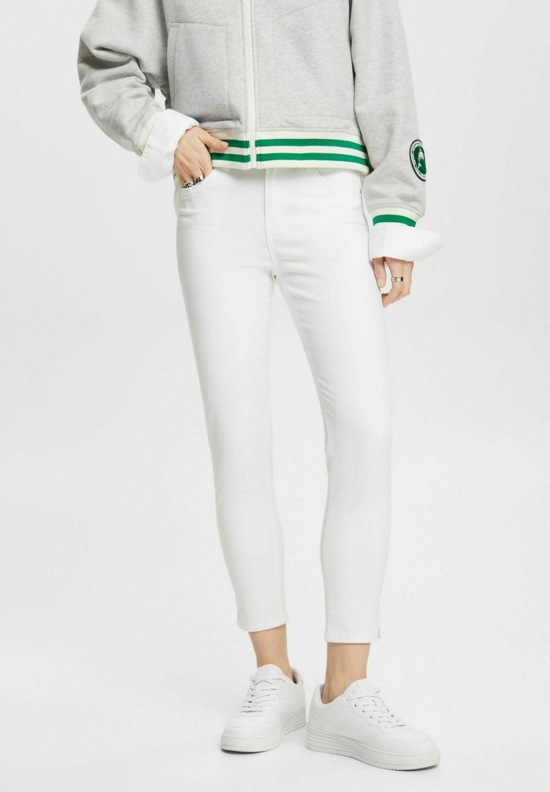Pantalons extensibles blancs avec une coupe ajustée, dotés d'une taille haute et de fermetures éclair discrètes aux chevilles, associés à une veste zippée grise.
