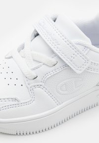 Białe skórzane sneakersy z perforowanymi detalami, płaskimi sznurowadłami i paskiem na rzep. Zawierają wytłoczone logo i teksturowaną gumową podeszwę.