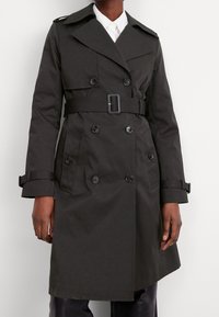 Mörkgrå trenchcoat i vattenavvisande tyg, med dubbelknäppt design, stora knappar och ett bälte i midjan.