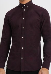 Muž na sobě má přiléhavou košili s dlouhým rukávem, kterou zdobí tmavě červený a černý malý kostkovaný vzor.