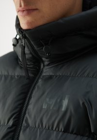 Black padded jacket featuring a high collar, zipper closure, and textured fabric. Subtle logo printed on the chest, adjustable toggles on the collar.