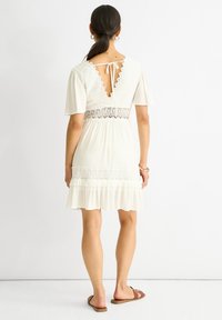 Robe blanche avec des accents en dentelle, manches courtes en forme de cloche et taille cintrée. Présente un dos en trou de serrure et un ourlet à volants en tiers. Portée avec des sandales marron.