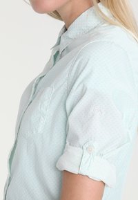 La camicia blu chiaro ha un sottile motivo a pois, una tasca sul petto e maniche arrotolate con dettagli a bottoni.