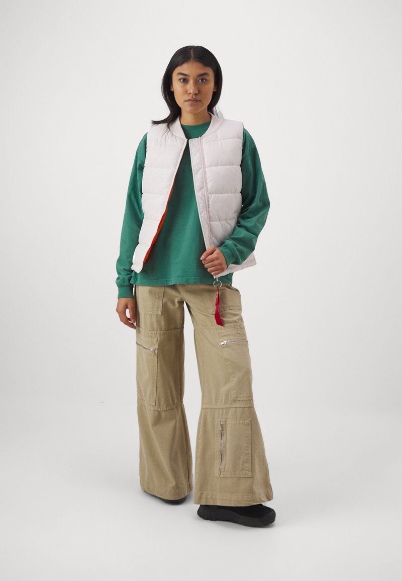 Wattierte Weste in Hellrosa mit Reißverschluss, getragen über einem grünen Langarmhemd und beigefarbenen, weit geschnittenen Cargo-Hosen mit mehreren Taschen.