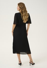 Melna plīsēta midi kleita ar caurspīdīgām plīvuru piedurknēm. Mīksta auduma, plūstoša tekstūra un pieguļošs jostas gabals. Melni slingback apavi noslēdz izskatu.