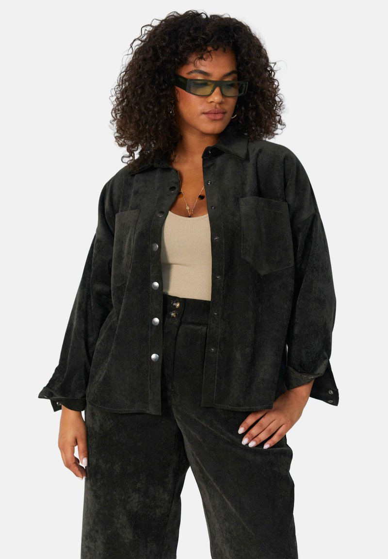Zwarte suède-achtige oversized shirt met knoopsluiting, twee voorzakken en opgerolde mouwen, gecombineerd met bijpassende broek.