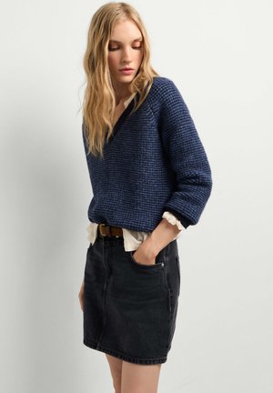Donna con lunghi capelli biondi che indossa un maglione blu lavorato, una camicetta bianca e una gonna di denim nera con le mani nelle tasche, guarda verso il basso.