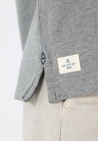 T-shirt en coton gris avec une étiquette cousue portant la mention "PAS JUSTE UN BR" et un emblème décoratif près de l'ourlet, finition en tissu texturé.