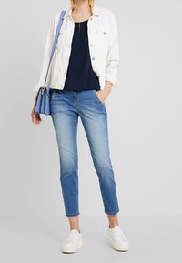 Weiße Denimjacke über einem marineblauen Oberteil, blaue Handtasche, hellblaue Jeans und weiße Sneakers. Bemerkenswerte Details sind ein Reißverschluss und Fronttaschen.
