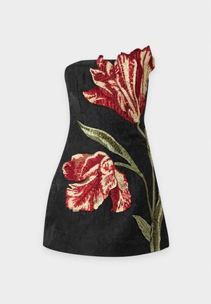 Femme portant une mini-robe noire bustier avec une grande broderie florale rouge et verte, associée à des sandales noires à talons hauts et des bracelets argentés.
