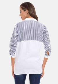 Das Hemd hat einen gestreiften Rücken in Marineblau und Weiß, eine einfarbige weiße Vorderseite, lange Ärmel mit Knopfdetails und einen lockeren Schnitt.