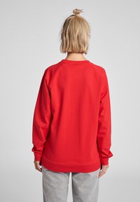 Felpa rossa con maniche raglan lunghe. Texture liscia e scollatura arrotondata. Abbinata a pantaloni della tuta grigi, vista di spalle.