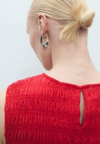 Haut smocké rouge avec un dos en trou de serrure ; associé à des boucles d'oreilles abstraites en argent qui reflètent la lumière. Texture lisse et design ajusté.