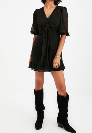 Vrouw die een zwarte mini-jurk met textuur draagt, met pofmouwen, een strikdetail aan de voorkant, een gerafelde zoom, een gouden kettingarmband en zwarte kniehoge laarzen.