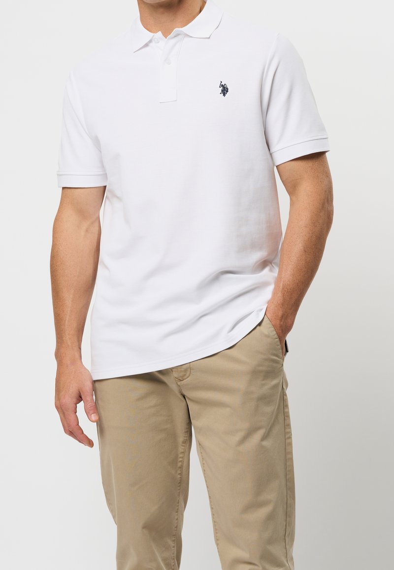 Homme portant un polo blanc à manches courtes avec un petit logo noir et un pantalon beige, une main dans la poche.