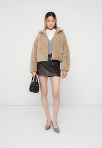 Kvinde iført en beige fuzzy jakke, hvid skjorte, grå strikvest, sort læder mini-nederdel og holder en sort håndtaske.
