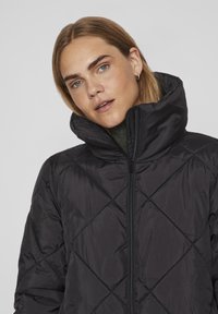 Črna prešita puhovka z visokim ovratnikom in zadrgo. Tkanina je gladka s rahlim sijajem, vsebuje diagonalne šive.