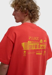 Persona joven con cabello rizado que lleva una camiseta roja con un gráfico amarillo de un estadio y texto en la espalda.