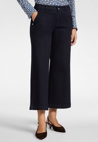 Donna con pantaloni cropped a gamba larga scuri, top a maniche lunghe con motivo, e scarpe slingback scure a tacco basso, in piedi con le mani nelle tasche.