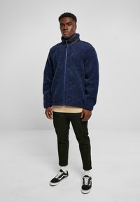 Marineblaue Fleecejacke mit hohem Kragen und Reißverschluss; kombiniert mit einem beigen Hemd, schwarzen Hosen und schwarzen Sneakern mit weißen Akzenten.