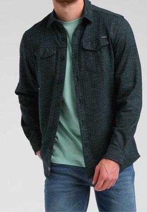 Mann trägt ein dunkles, strukturiertes Hemd mit Knöpfen über einem hellgrünen T-Shirt und blauen Jeans, steht mit einer Hand in der Tasche.