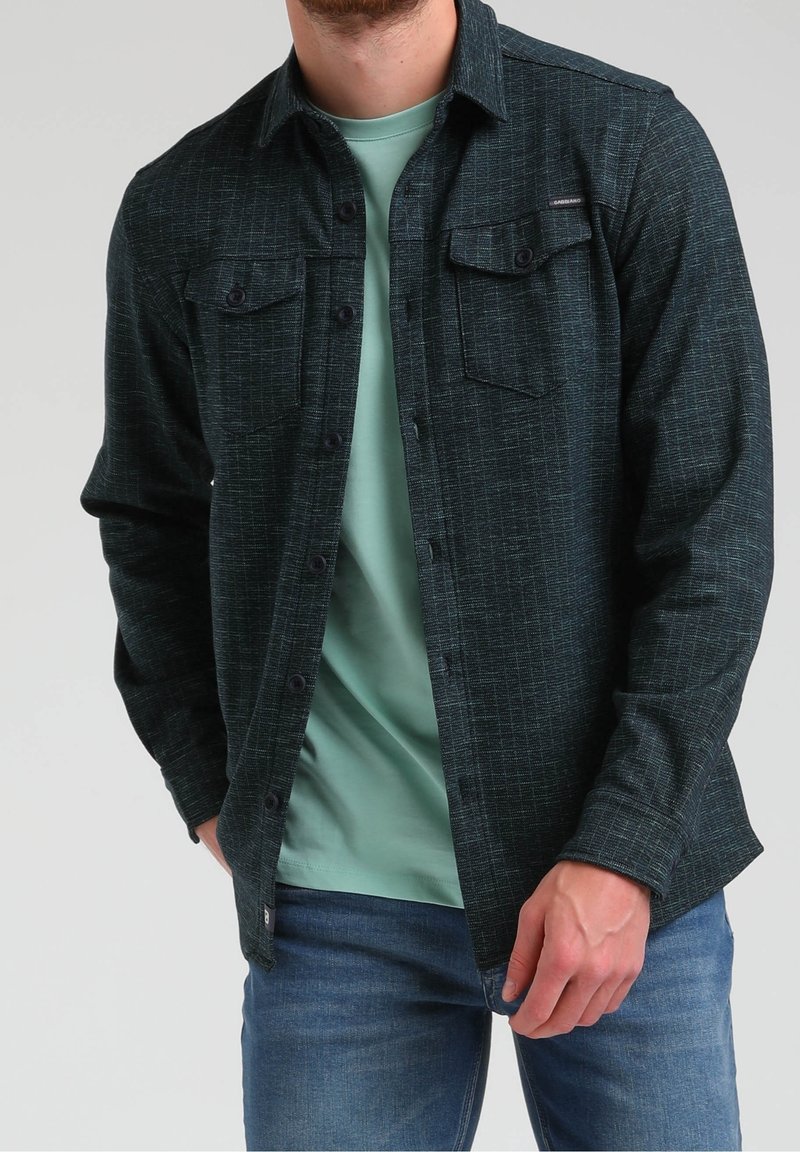 Homme portant une chemise boutonnée texturée foncée par-dessus un t-shirt vert clair et un jean bleu, debout avec une main dans la poche.