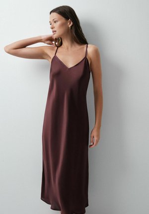 Femme portant une robe nuisette en satin violet foncé sans manches, debout contre un mur gris clair, une main touchant son cou.