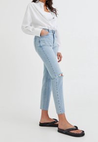 Jeans de cintura alta de color azul claro con detalle desgastado en las rodillas, combinados con una camisa blanca de botones y chanclas negras. Tejido de algodón, corte entallado.