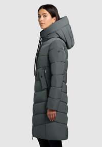 Vrouw in een lange donkergrijze gewatteerde winterjas met een grote capuchon, een ritszak en een hoge kraag, staande in zijaanzicht.