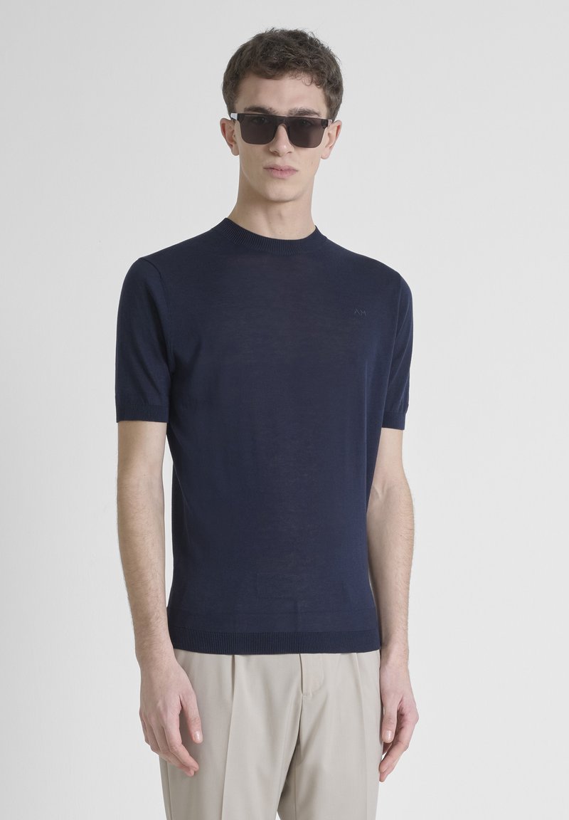 Jersey de manga corta azul marino con cuello redondo, que presenta un sutil detalle de logo y puños acanalados, combinado con pantalones beige.