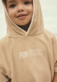 Beige håndklæde poncho med hætte, der har korte ærmer og en blød tekstur. "FUNtastic" er broderet i hvidt på forsiden.