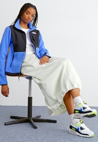 Tânără cu părul împletit, așezată pe un scaun cu rotile, purtând o jachetă din polar albastră și neagră, o fustă albă și adidași albi cu detalii albastre și verzi neon.