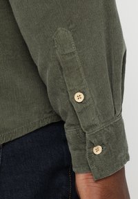 Camicia in corduroy verde oliva con maniche, dotata di due bottoni in legno, caratterizzata da una texture a coste e dettagli con orlo arrotolato.