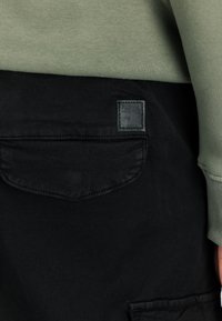 Černé kalhoty se zadní kapsou s obdélníkovou šedou nášivkou s vyraženým nápisem "ALPHA INDUSTRIES". Horní část je vyrobena z olivově zelené látky.