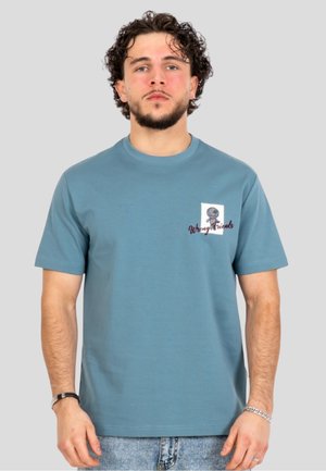 Mand med krøllet hår og skæg iført en blå t-shirt med lille grafik og tekst "Wrong Friends" på venstre bryst, stående mod en grå baggrund.