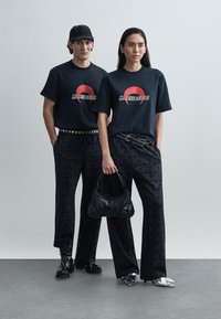 Black t-shirts with red graphic text, loose black patterned trousers, black handbag, and shiny silver shoes. Both individuals stand side by side.