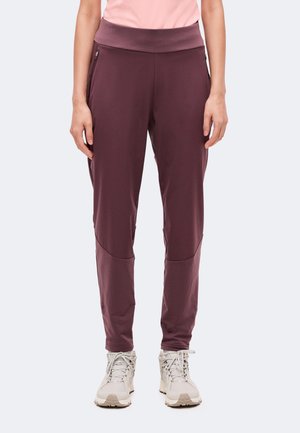 Personne portant un pantalon de sport bordeaux avec des poches zippées et des chaussures de course beiges sur un fond clair uni.