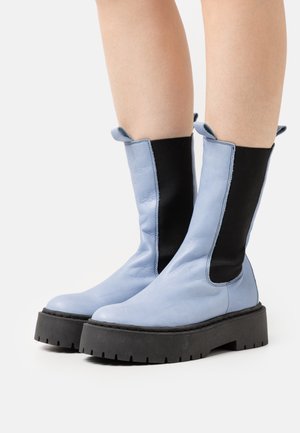 Botas de tobillo de cuero azul claro con paneles laterales de elastano negro, suelas de goma negra gruesa con un patrón de banda de rodadura resistente.