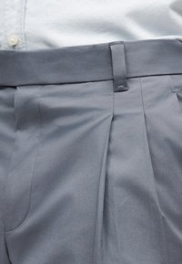 Ανοιχτού μπλε παντελόνι με θηλιές για ζώνη και λεπτομέρεια με πιέτες, φορεμένο με λευκό πουκάμισο που έχει μπει μέσα και κουμπωμένο στη μέση.