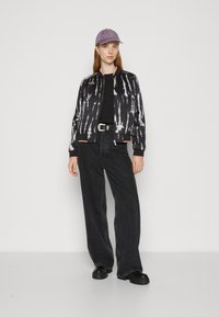 Veste bomber noire avec un motif tie-dye, accents roses, associée à un top noir, un jean noir ample et des chaussures noires massives. Casquette violette.