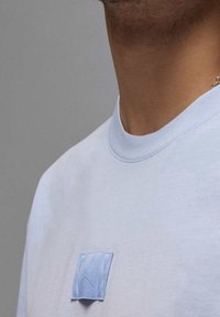 Světle modrý svetr s kulatým výstřihem, zdobený malou modrou nášivkou. Materiál má jemný a hladký povrch.