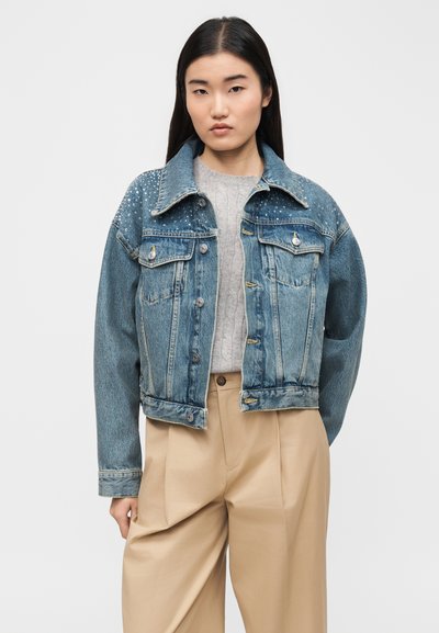 Νεαρή γυναίκα που φοράει μπλε τζιν μπουφάν με στρας, γκρι πλεκτό πουλόβερ και μπεζ ψηλόμεσο παντελόνι, στέκεται μπροστά σε απλό φόντο.