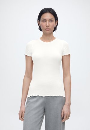 Frau mit kurzen schwarzen Haaren, die ein eng anliegendes weißes Kurzarmshirt und graue Hosen trägt, steht vor einem schlichten hellen Hintergrund.