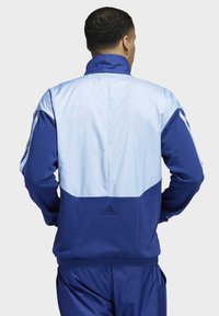 Veste bleue avec des accents bleu clair, col zippé et poignets élastiques. Présente le logo Adidas au dos avec une finition texturée.