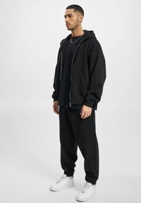 Sort hættetrøje med lynlås og forlommer, parret med en sort crewneck t-shirt og sorte joggers. Accenteret med hvide sneakers.