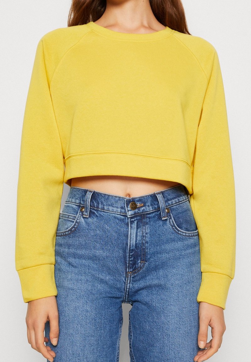 Femme portant un sweat-shirt court jaune vif à manches longues et un jean bleu taille haute, montrant le ventre et avec une braguette à boutons.